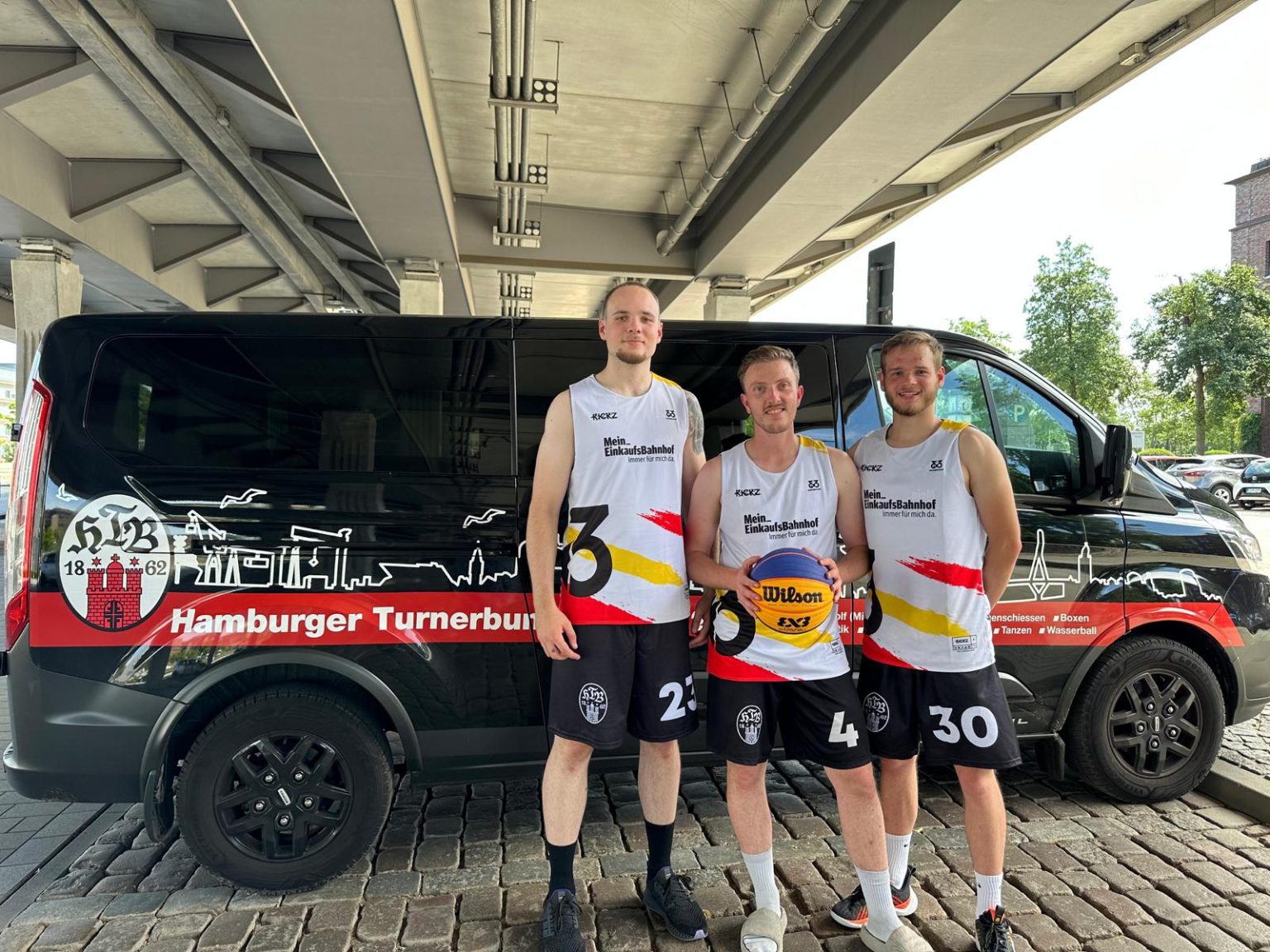 Drei HTB62-Spieler in 3x3-Trikots mit einem Basketball vor dem Vereinsbus
